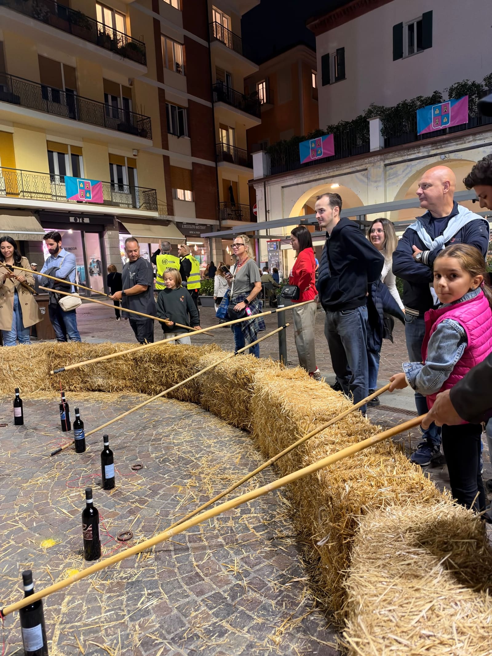 eccoci a un gioco dei Brichett in Piazza Pertinace: con una canna lunga si prova a infilare un anello al collo delle bottiglie, oltre le balle di paglia