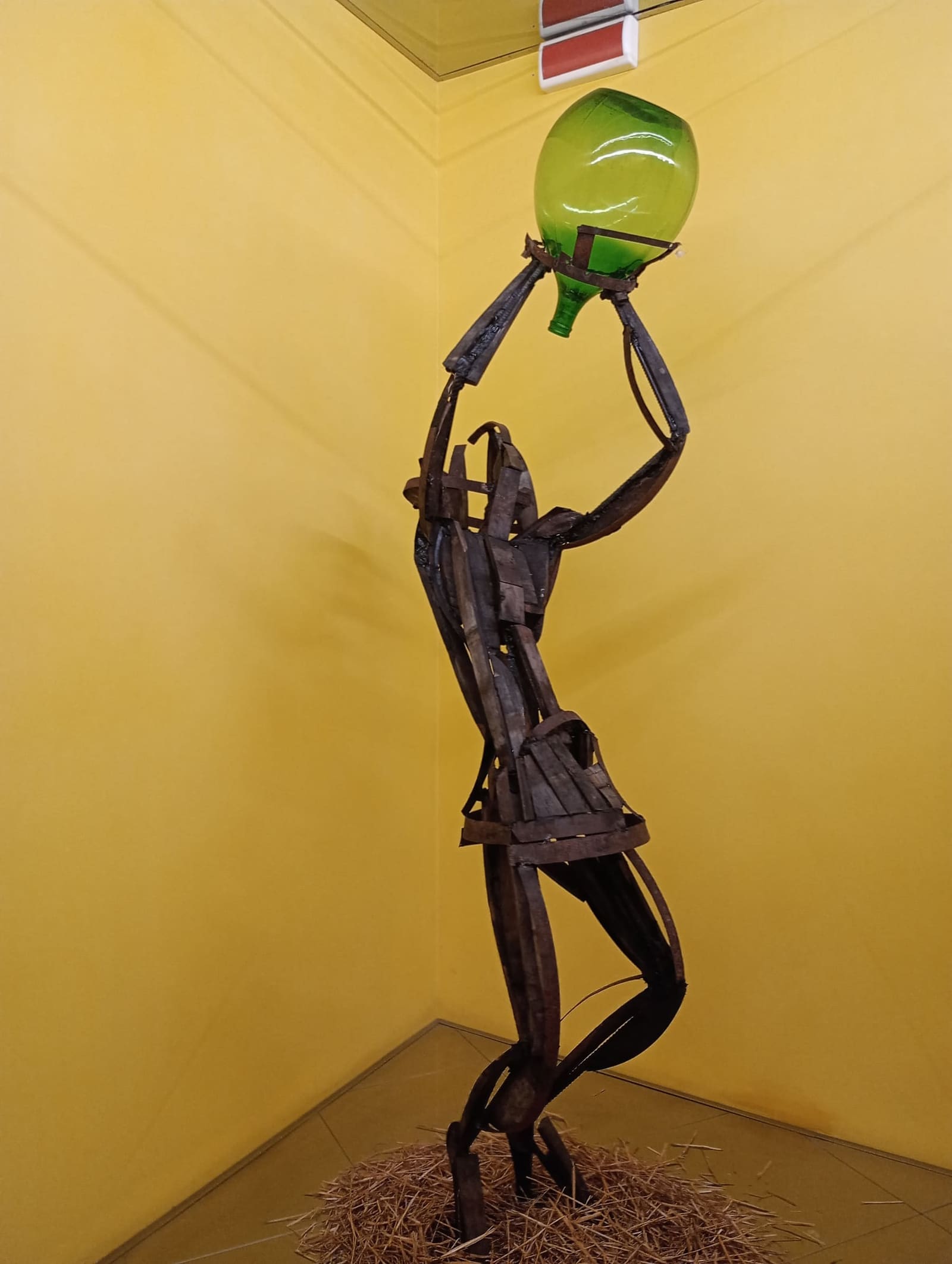 una figura di ferro che solleva una damigiana verde, in una sala del WiMu