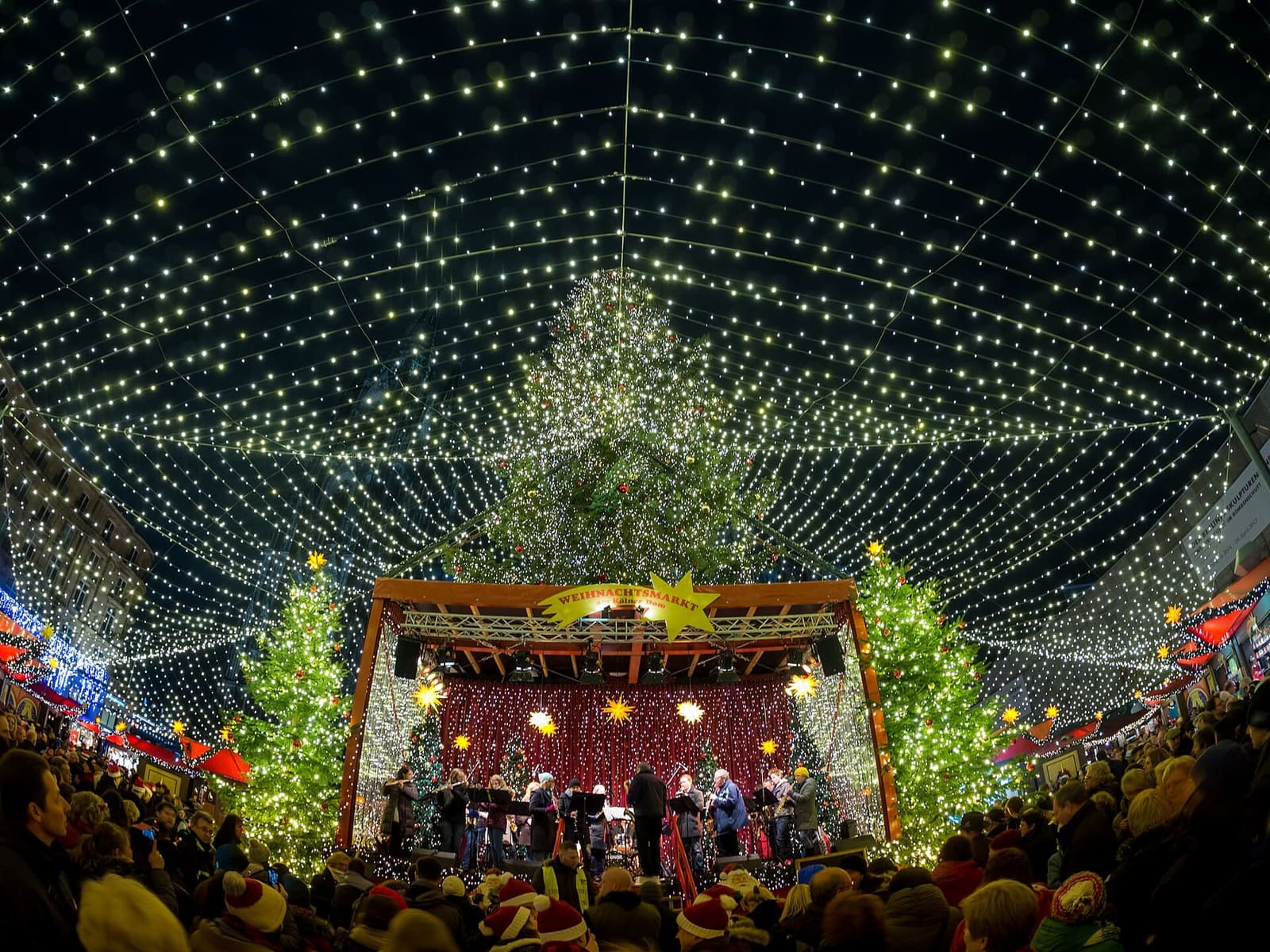 il palco del mercatino davanti al Duomo sotto il tetto di lucine, con l'albero e la folla della sera