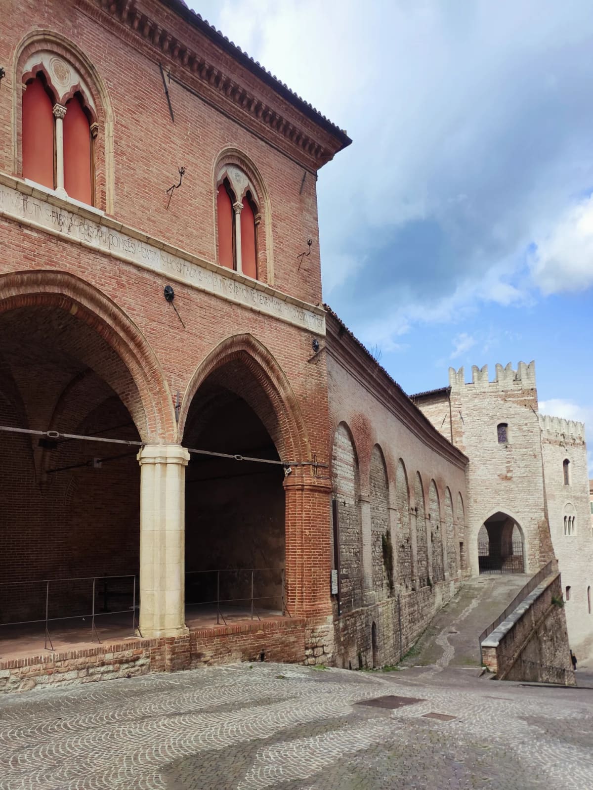 le arcate gotiche dell'ospedale con l'iscrizione latina incisa nella fascia di pietra, e in fondo la torre merlata