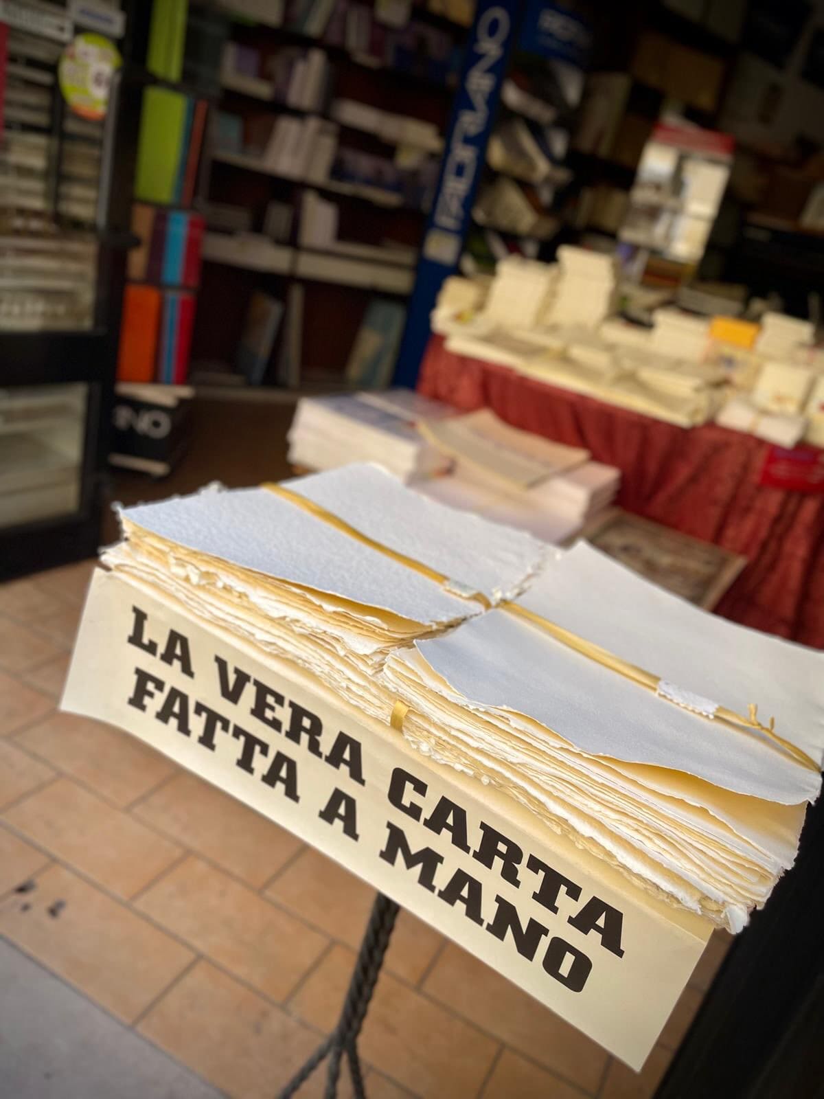 il cartello che dice "la vera carta fatta a mano" appoggiato a una risma di fogli dai bordi sfrangiati, sulla porta della cartoleria