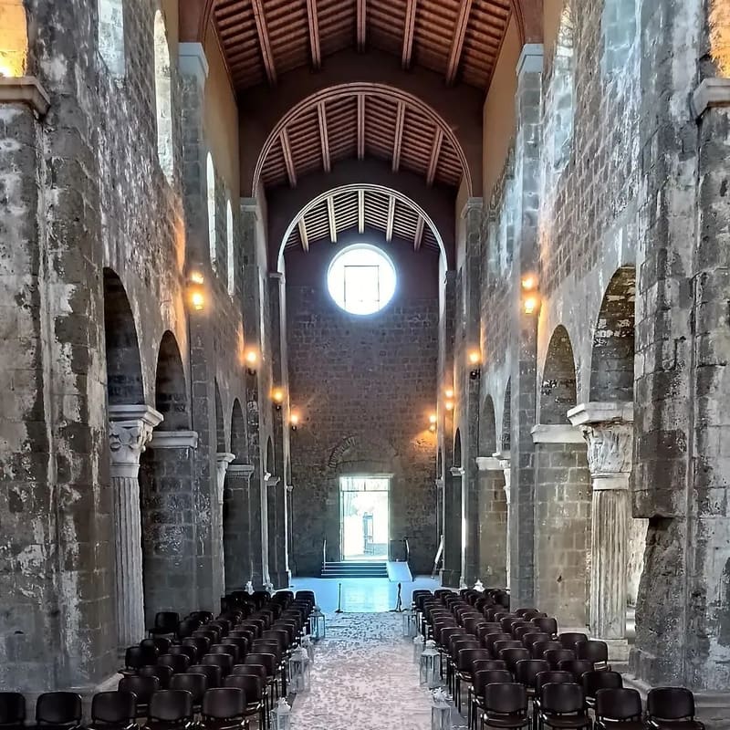 dentro l'abbazia: tre navate di pietra nuda, le capriate di legno a vista, il rosone in fondo, e le sedie in fila per una funzione