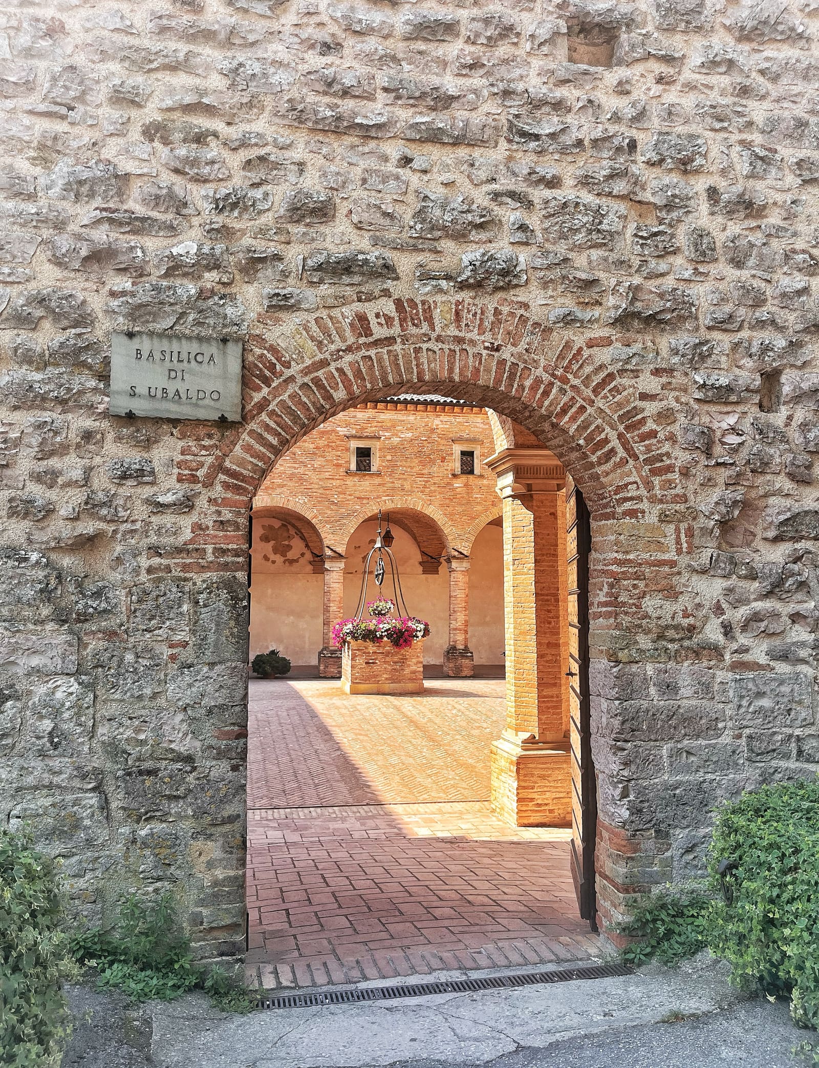 il portale con la lapide della basilica di Sant'Ubaldo, e dietro il chiostro di mattoni con il pozzo fiorito