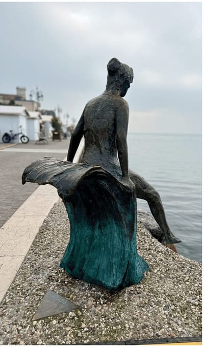 la statua di bronzo di una donna seduta in riva al lago, di spalle, con il panneggio verde di patina che scende sul basamento