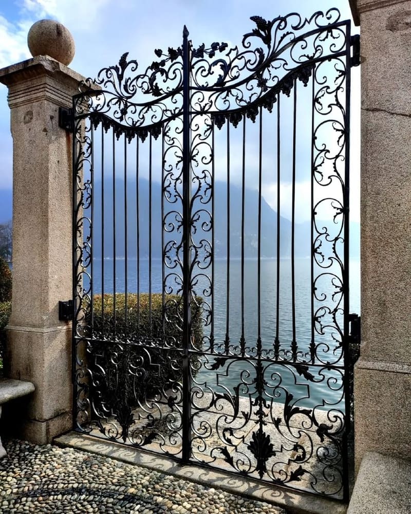 un cancello in ferro battuto aperto sul lago, con il selciato a ciottoli e le montagne nella foschia