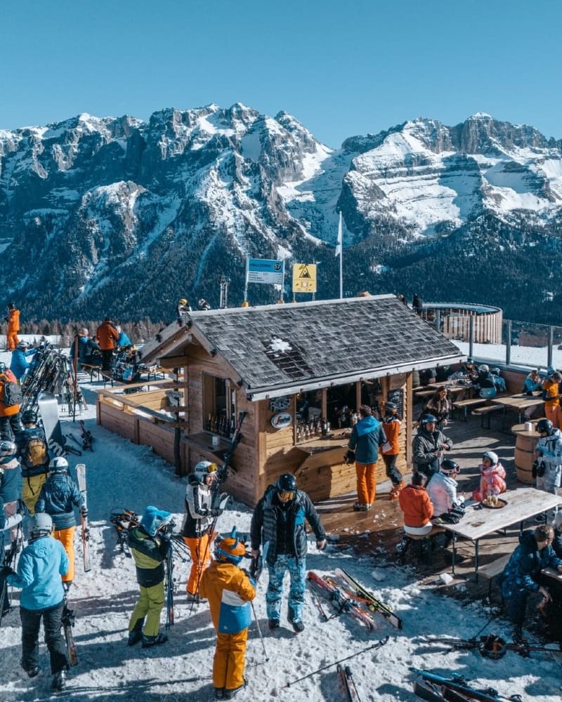 un chiosco di legno sulle piste in quota, sci appoggiati fuori e la catena del Brenta di fronte