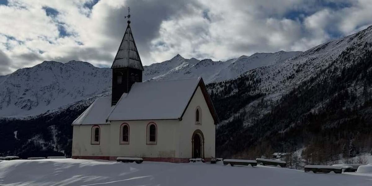 una chiesetta bianca con il campanile aguzzo in legno, panchine sepolte dalla neve e le cime alle spalle