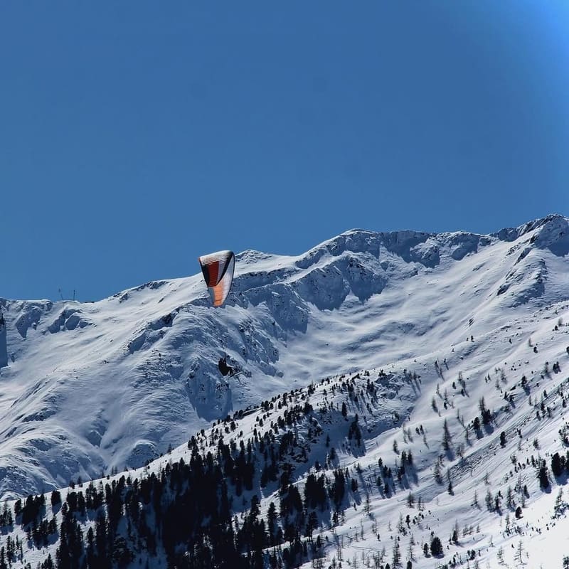 un parapendio sopra la cresta innevata, nel cielo blu sopra il bosco di larici
