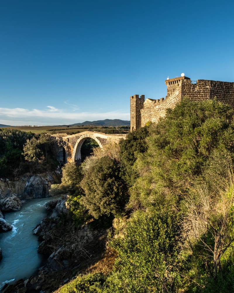 il ponte dell'Abbadia visto da valle, con l'arcata romana sopra le rocce e il Fiora che scorre pieno, e in alto a destra le mura merlate del castello