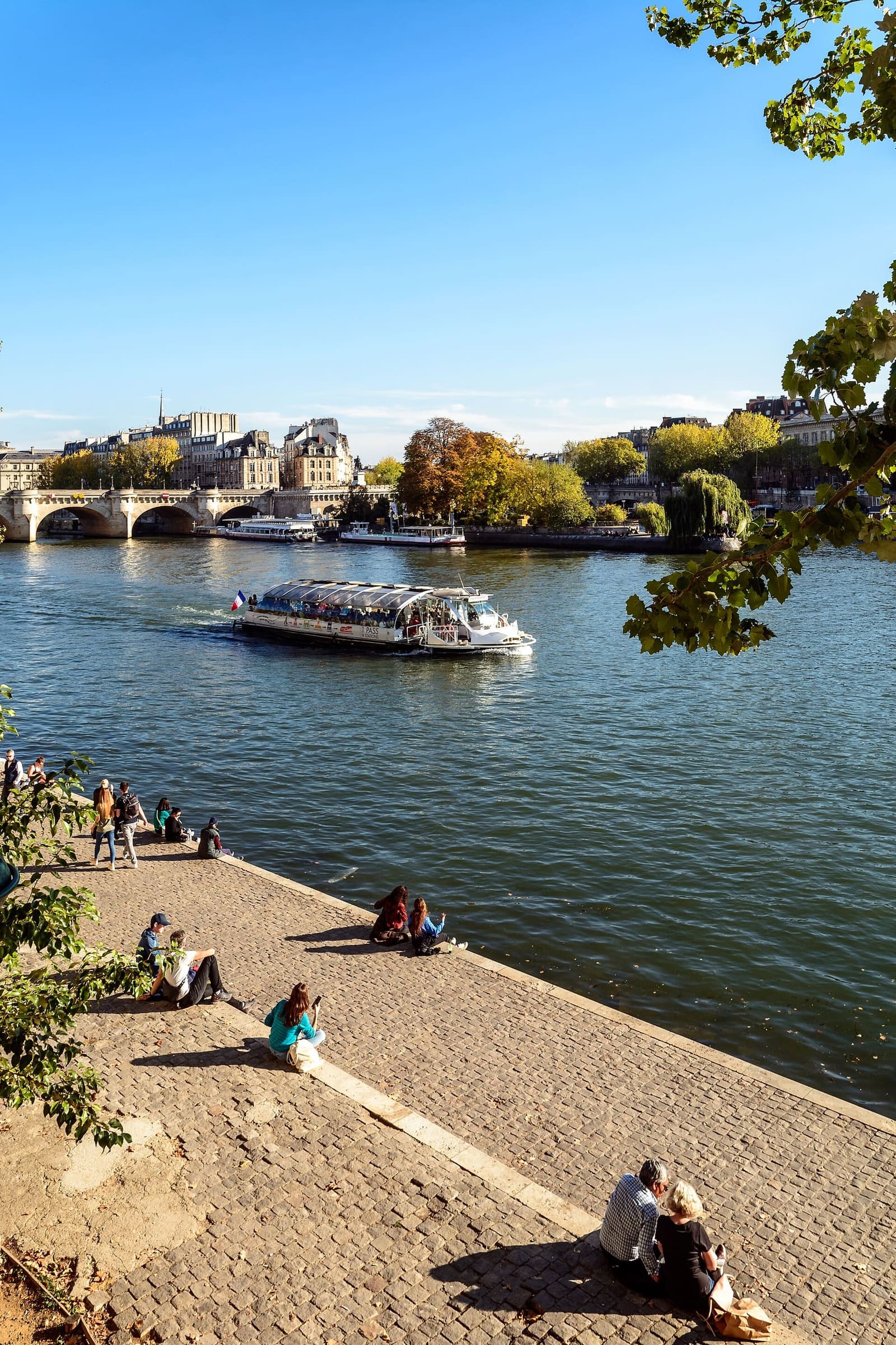 Menschen sitzen am Quai entlang der Seine, mit einem Boot und der Île de la Cité in der Ferne