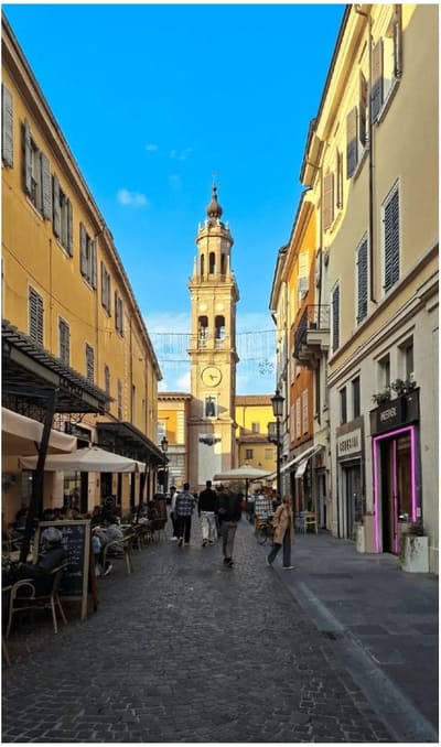 una via del centro con i tavolini fuori e un campanile con l'orologio in fondo