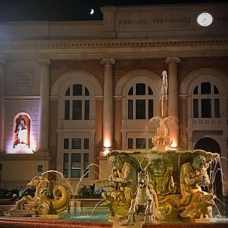 la fontana di Piazza del Popolo illuminata di notte, davanti al palazzo delle Poste