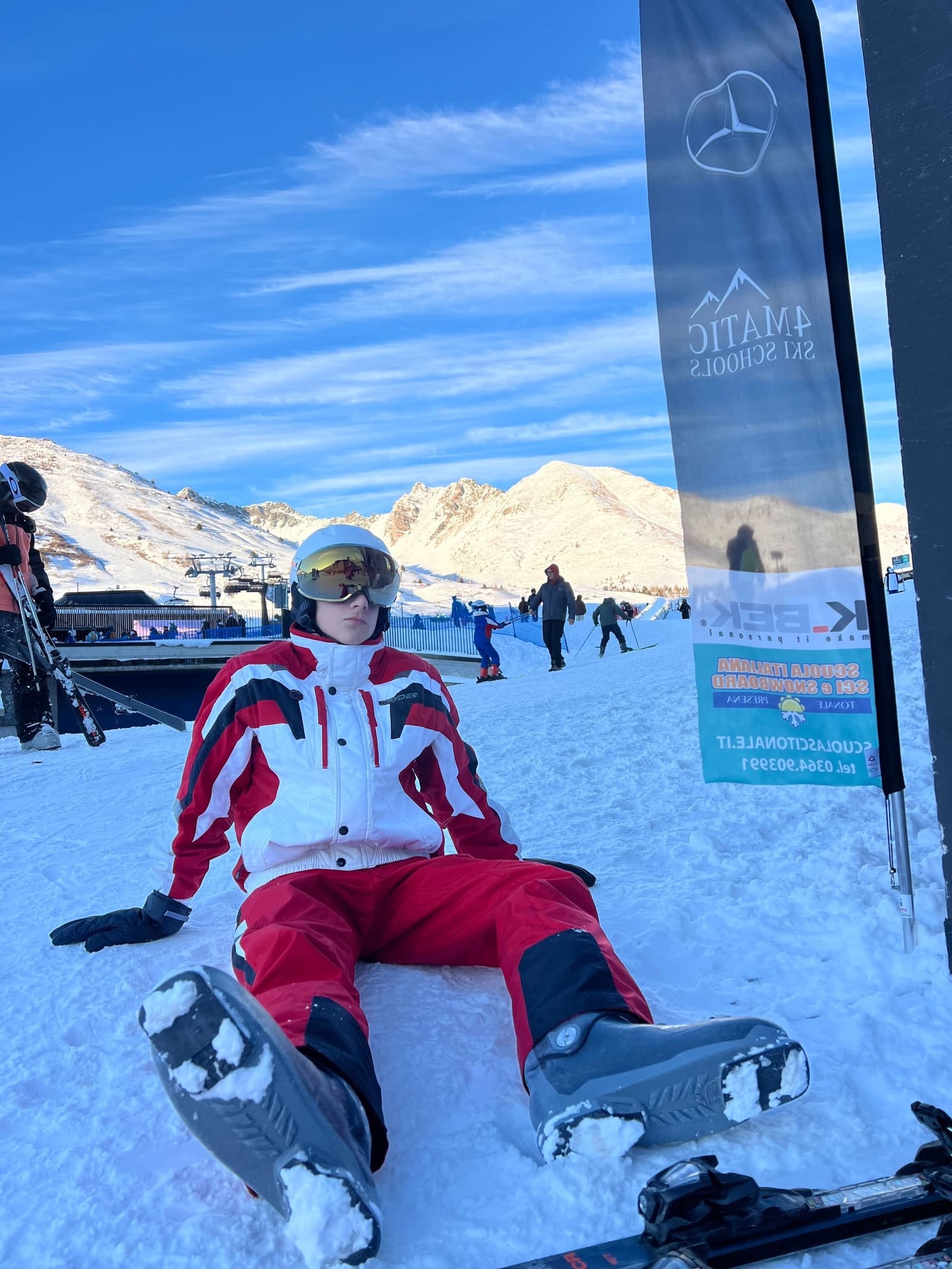 Gabriele seduto sulla neve davanti al gonfalone della scuola di sci, con le cime accese dal tramonto