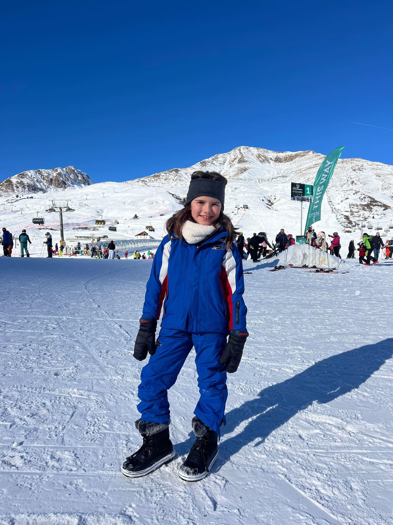 nostra figlia in tuta blu sulla neve, con le seggiovie e il cartello di partenza dietro