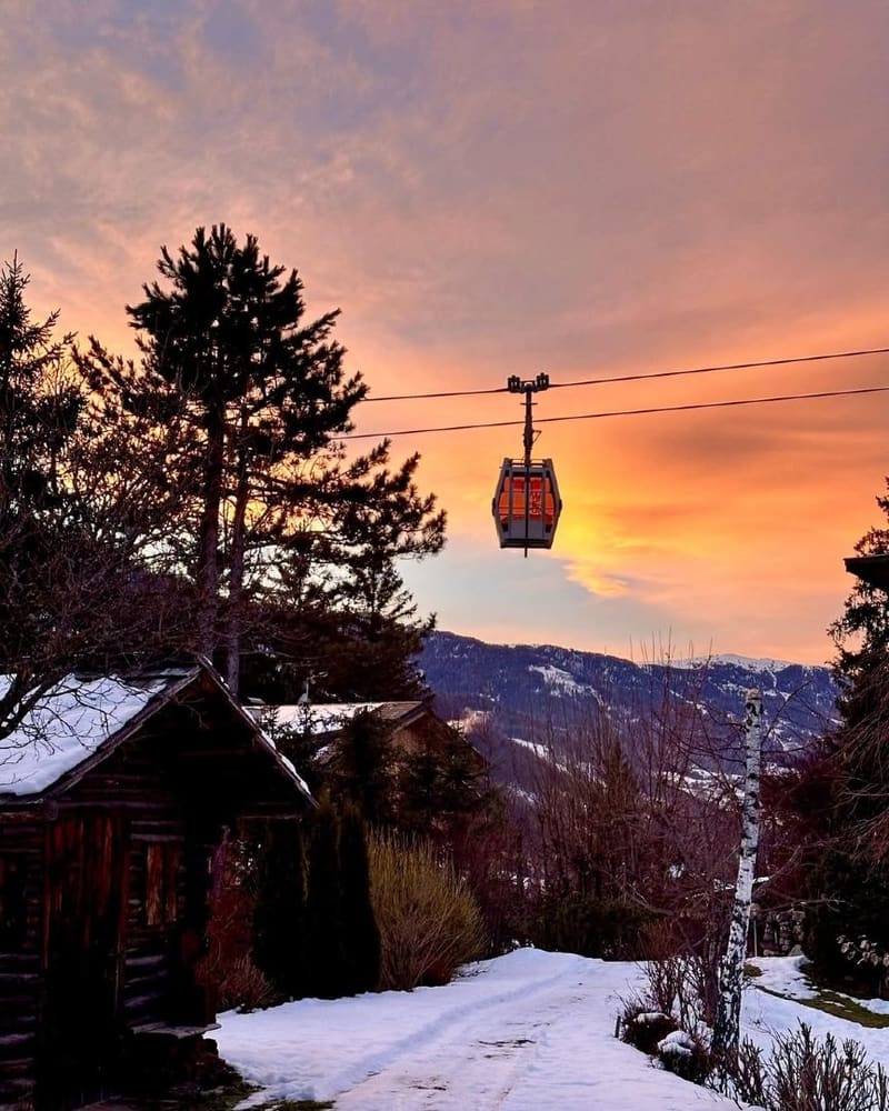 una cabina della funivia sospesa sul cielo arancione, sopra i tetti innevati