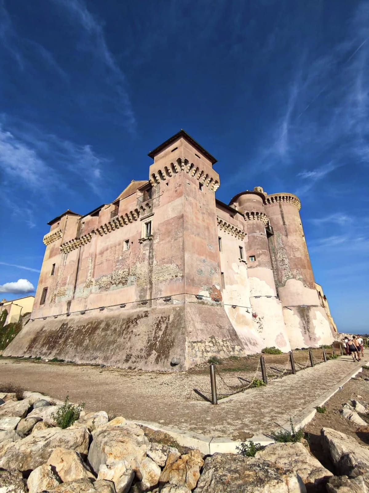 il castello dallo spigolo che guarda il mare, preso dal basso: la scarpa alla base, la fila dei beccatelli sotto il coronamento, la torre quadrata in mezzo e quella rotonda a destra, e davanti i blocchi bianchi della scogliera