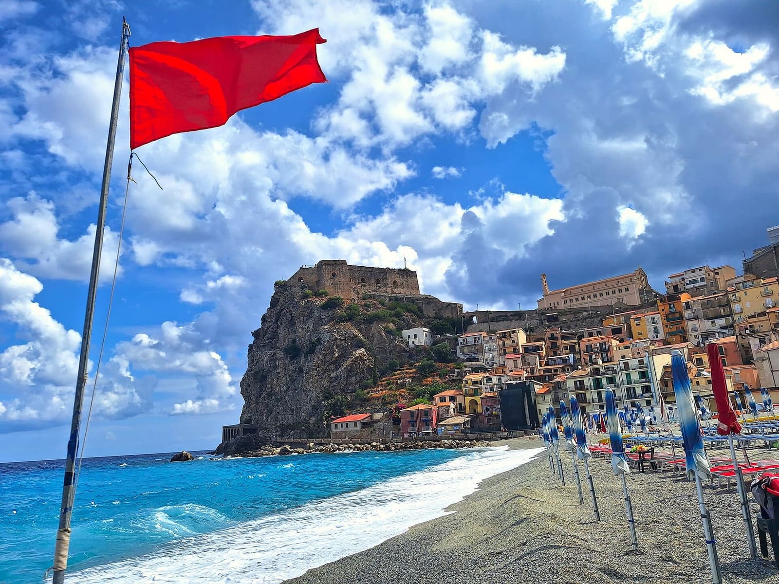the beach with the umbrellas closed and the red flag, and the rock in the distance