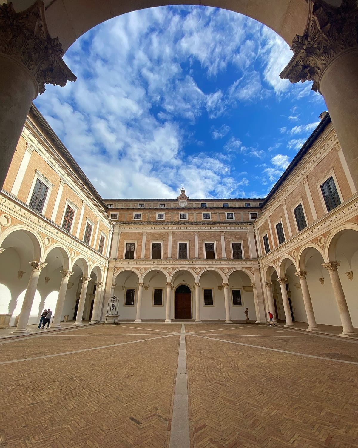 il cortile d'onore inquadrato da sotto un arco, con l'iscrizione latina che gira su tutti e quattro i lati e nomina Federico duca di Urbino