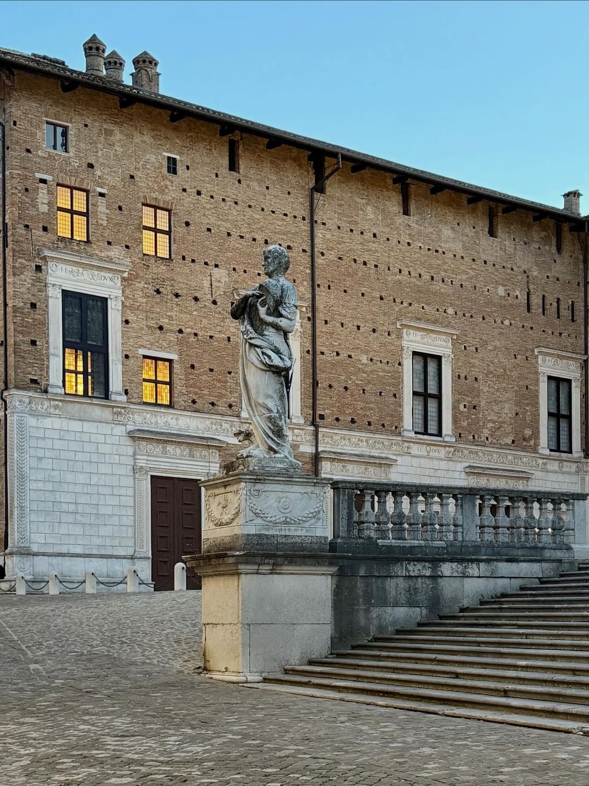 la statua di marmo sul piedistallo davanti alla facciata di mattoni del palazzo, con le finestre accese e i fregi che ripetono FE DVX