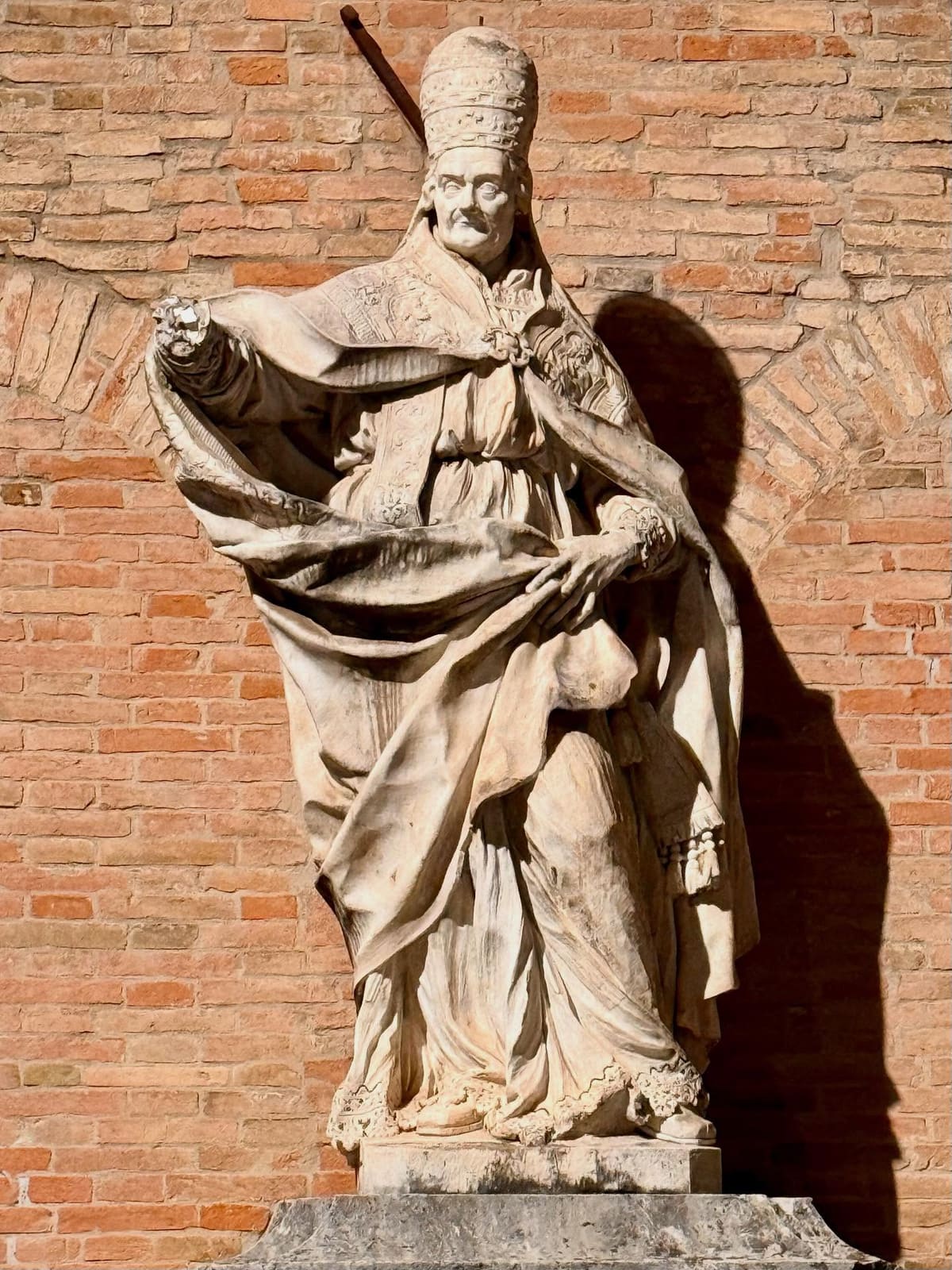 la statua di marmo di un papa in piviale e triregno, addossata al muro di mattoni, con la sua ombra netta accanto