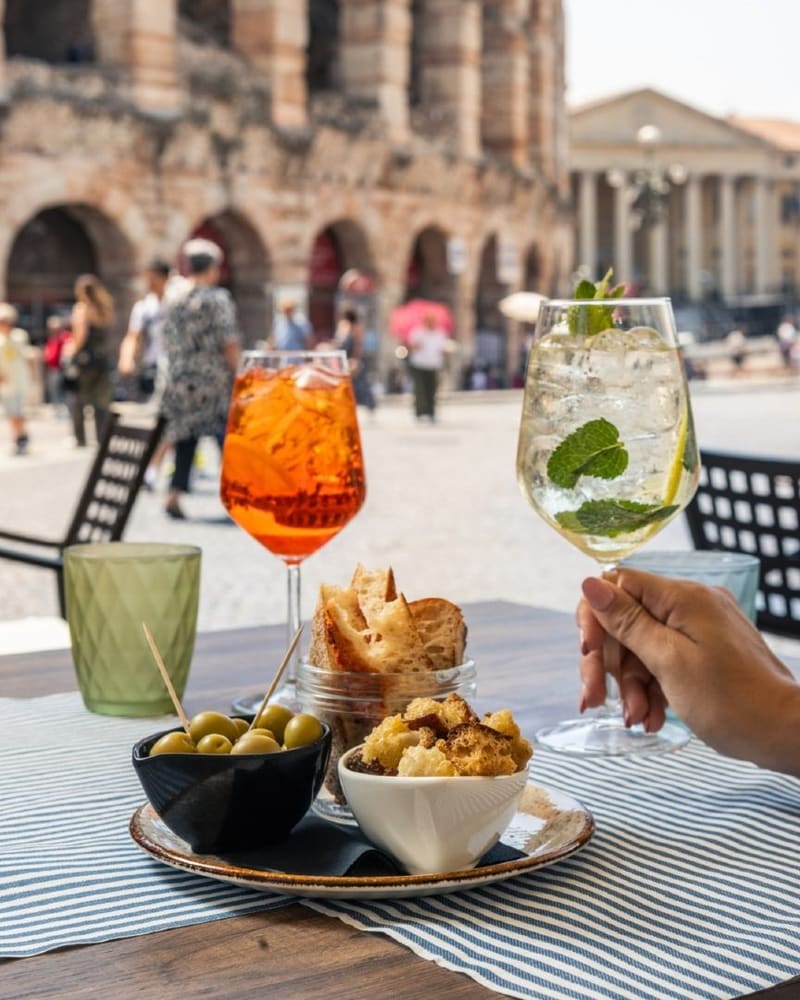 due calici e un piatto di olive e crostini su un tavolino, con le arcate dell'Arena sullo sfondo