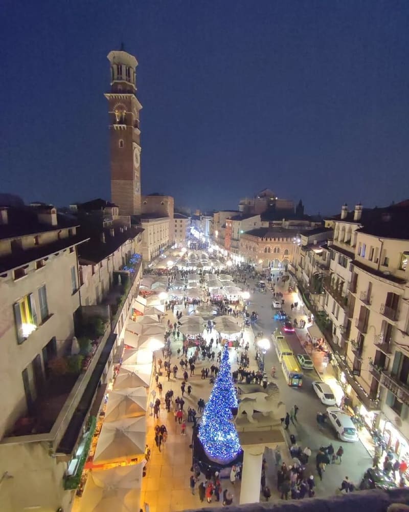 Piazza delle Erbe di sera dall'alto, i banchi bianchi del mercatino, l'albero di Natale illuminato e la Torre dei Lamberti